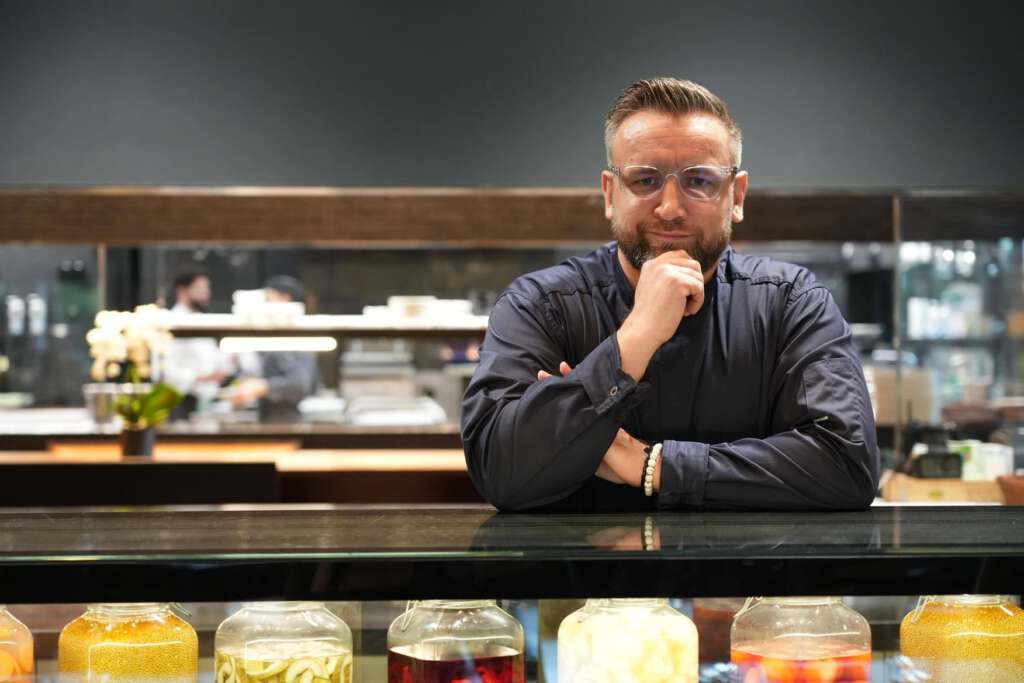 Ein Koch mit Brille steht hinter einer Theke mit Gläsern voller Konserven.