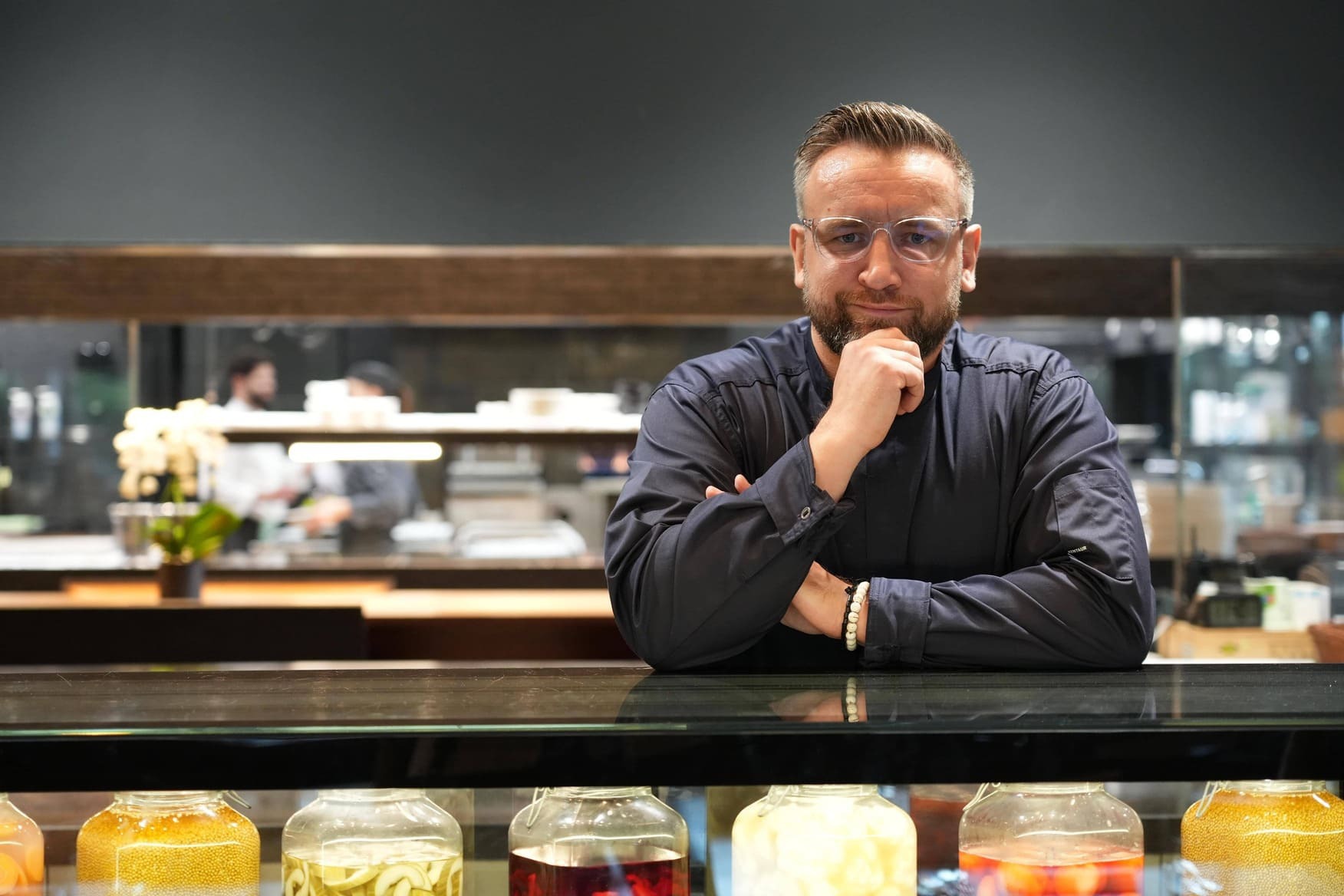 Ein Koch mit Brille steht hinter einer Theke mit Gläsern voller Konserven.