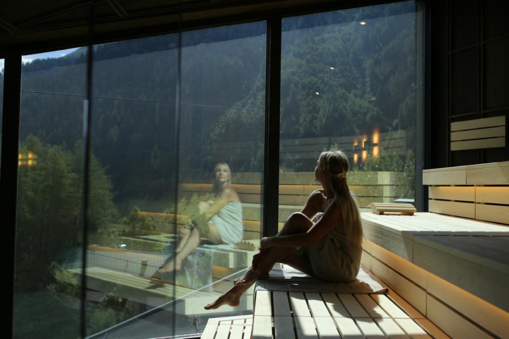 Eine Frau schaut aus einer Panorama-Sauna durch das Glas in die umliegende Berglandschaft und wird dabei mit von der Sonne angestrahlt.