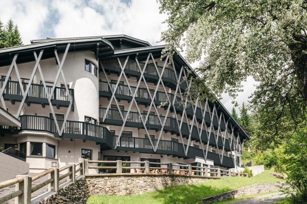 Ein mehrstöckiges Gebäude mit Holzbalkonen und Gitterdesign vor einer Kulisse aus Bäumen und einem bewölkten Himmel.