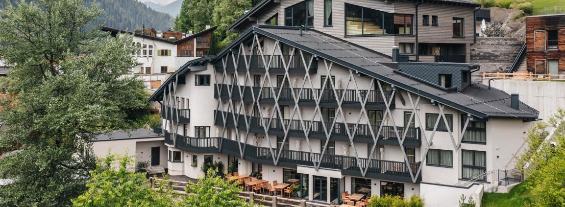 Ein großes, leicht spitz zulaufendes Gebäude in weiß mit dunkelgrünen Verstrebungen und Balkonen. Auf dem Dach befindet sich ein Wellnessbereich mit Saunen, darüber sind viele Nadelbäume zu sehen.