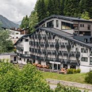 Ein großes, leicht spitz zulaufendes Gebäude in weiß mit dunkelgrünen Verstrebungen und Balkonen. Auf dem Dach befindet sich ein Wellnessbereich mit Saunen, darüber sind viele Nadelbäume zu sehen.