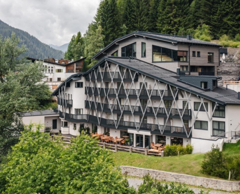 Ein großes, leicht spitz zulaufendes Gebäude in weiß mit dunkelgrünen Verstrebungen und Balkonen. Auf dem Dach befindet sich ein Wellnessbereich mit Saunen, darüber sind viele Nadelbäume zu sehen.