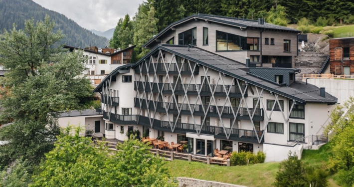 Ein großes, leicht spitz zulaufendes Gebäude in weiß mit dunkelgrünen Verstrebungen und Balkonen. Auf dem Dach befindet sich ein Wellnessbereich mit Saunen, darüber sind viele Nadelbäume zu sehen.