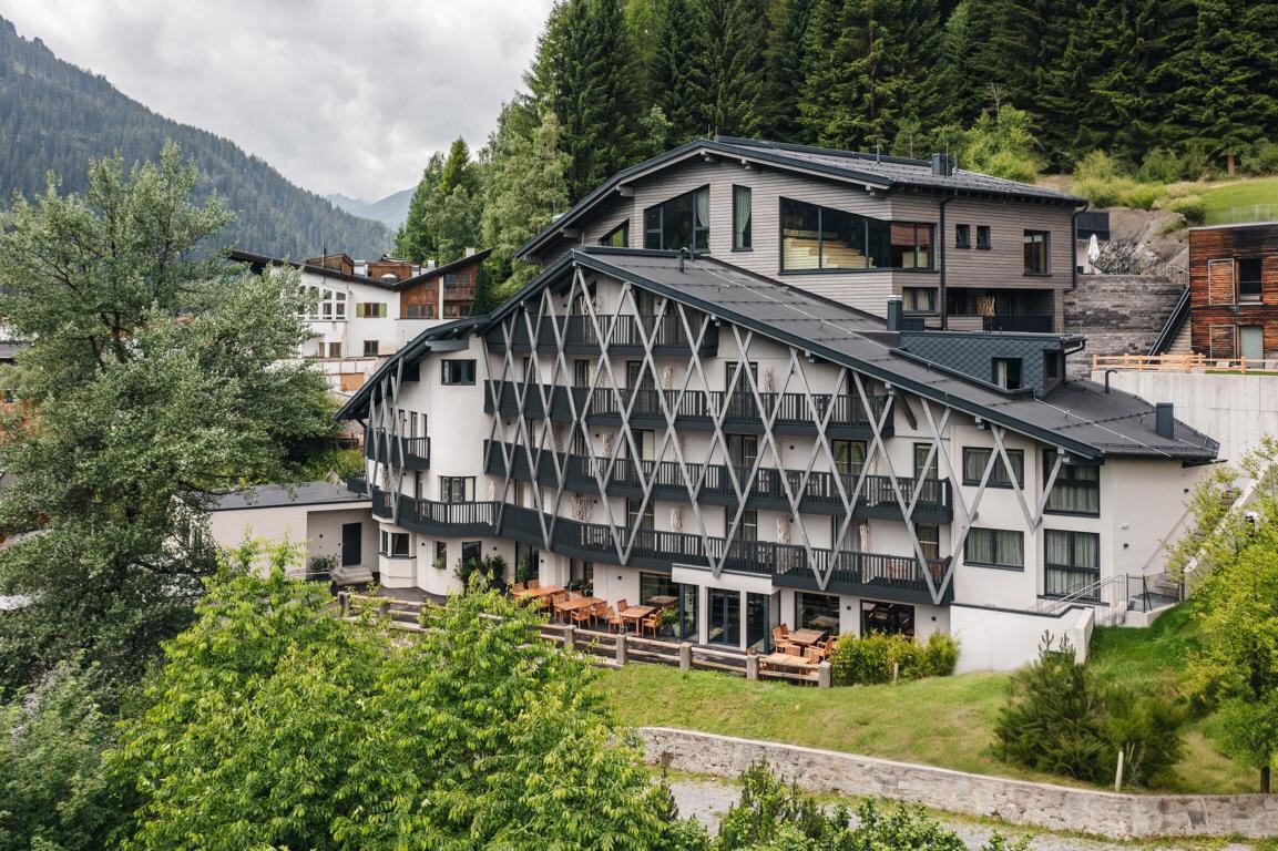 Ein modernes, mehrstöckiges Gebäude im Chalet-Stil mit geometrischen Fassadenelementen, umgeben von üppigem Grün und Bergen im Hintergrund.