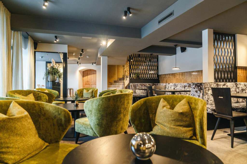 Ein modernes Interieur mit grünen Samtstühlen, schwarzen Tischen und dekorativen Metalltrennwänden. Der Raum ist gut beleuchtet und bietet eine Mischung aus Texturen und neutralen Tönen.