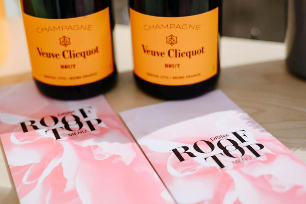 Zwei Flaschen Champagner Veuve Clicquot Brut werden neben zwei Getränkekarten mit rosa Blumenmuster auf einer Dachterrasse auf einer Holzoberfläche präsentiert.