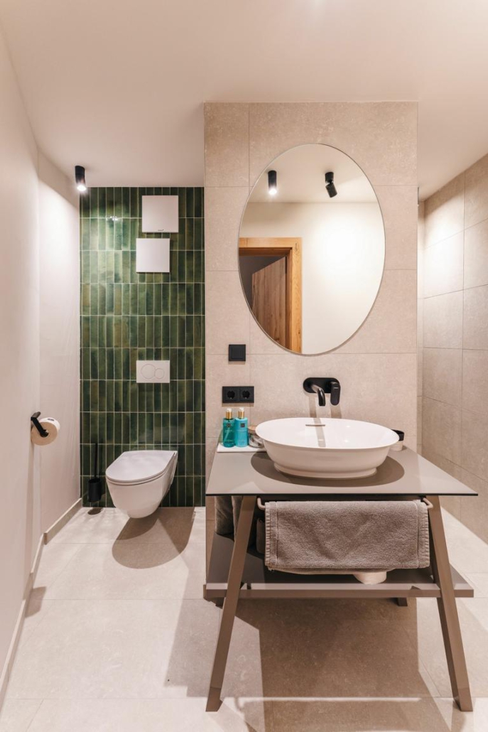 Blick in ein Badezimmer eines Hotels in St. Anton in Österreich, das von warmen Steintönen und minimalistischer Ausstattung geprägt ist. Vorne ist der Waschtisch zu sehen, links eine Toilette an einer grün gekachelten Wand.