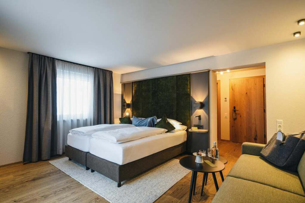 Ein Hotelzimmer mit einem großen Bett, einem grünen Kopfteil, einem Sofa, Holzboden und einem kleinen runden Tisch mit Wasserflaschen. Im Hintergrund gibt es ein Fenster mit Vorhängen und eine Holztür.