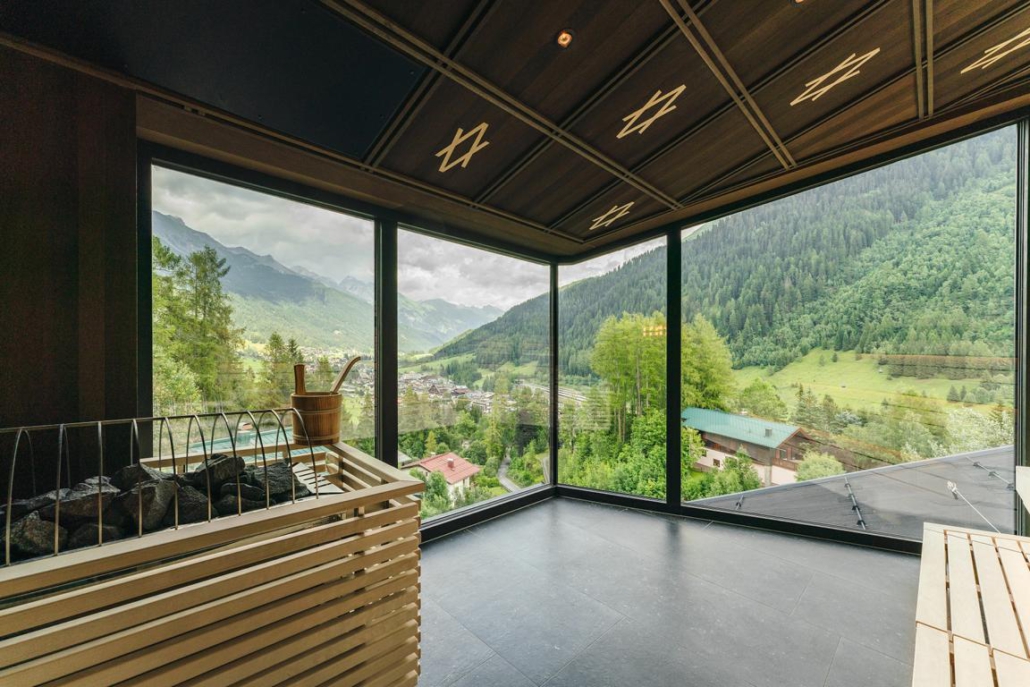 Blick aus der Panorama-Sauna eines Hotels in St. Anton am Arlberg in Österreich. Links ist ein moderner Saunaofen mit Holzbeschlag zu sehen, rechts daneben bieten große Fensterfronten einen weiten Panoramablick auf das darunter liegende Tal.