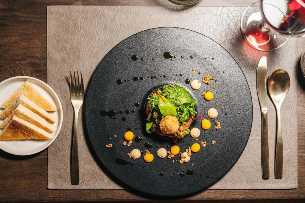 Ein Gourmetgericht auf einem schwarzen Teller mit Gemüse, etwas Gebäck und Soßen. Besteck an den Seiten, geröstetes Brot auf einem Beilagenteller und daneben ein Glas mit roter Flüssigkeit.