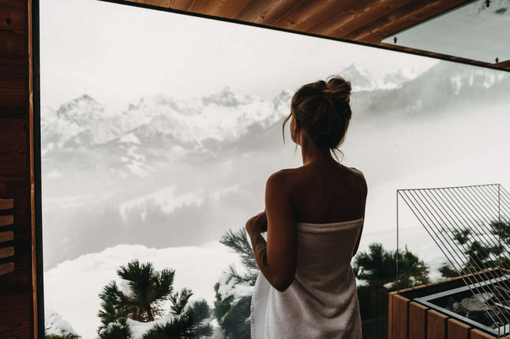 Eine Frau steht in ein Handtuch eingehüllt und blickt von einer Terrasse auf eine schneebedeckte Berglandschaft.
