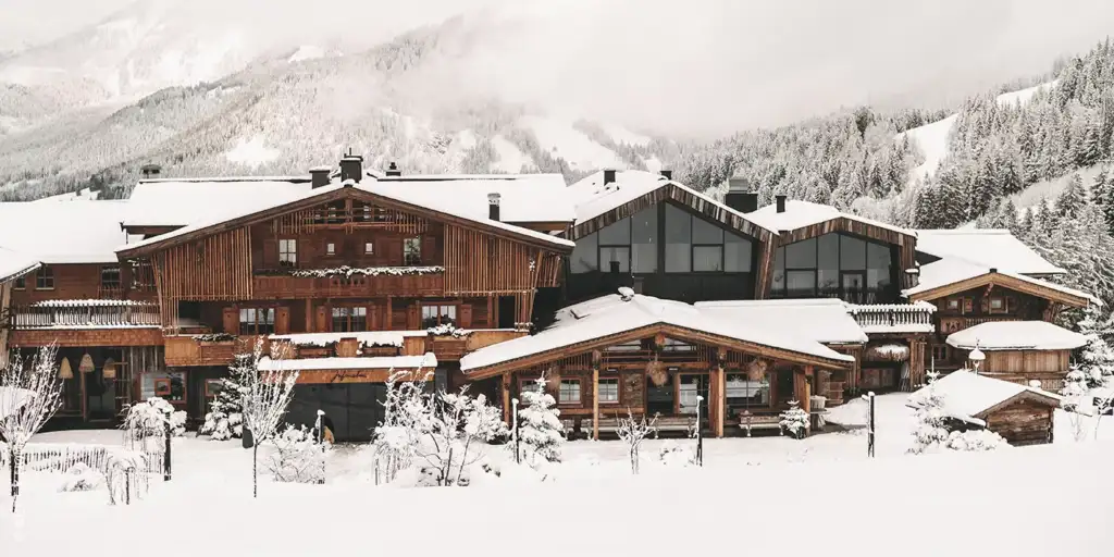 Schneebedeckte Holzgebäude im Chalet-Stil vor der Kulisse schneebedeckter Bäume und Berge unter einem bewölkten Himmel.