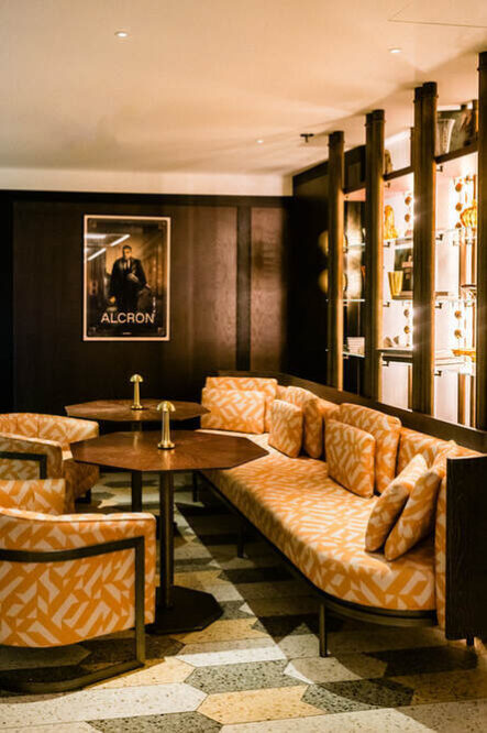 Eine gemütliche Lounge mit gemusterten orangefarbenen Sitzgelegenheiten, einem runden Tisch in der Mitte und einer Wand mit beleuchteten Regalen bestimmt die Atmosphäre. Ein gerahmtes „Alcron“-Poster ziert die holzgetäfelte Wand und erinnert an die schicke Eleganz der Alcron Bar Prag.