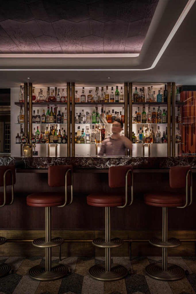 Ein Barkeeper steht hinter dem schwach beleuchteten Ambiente der Alcron Bar in Prag, in der Reihen von Schnapsflaschen sanft glänzen. Vier Barhocker säumen die Theke und laden die Gäste ein, in diesem gemütlichen Rückzugsort einen Drink zu genießen.