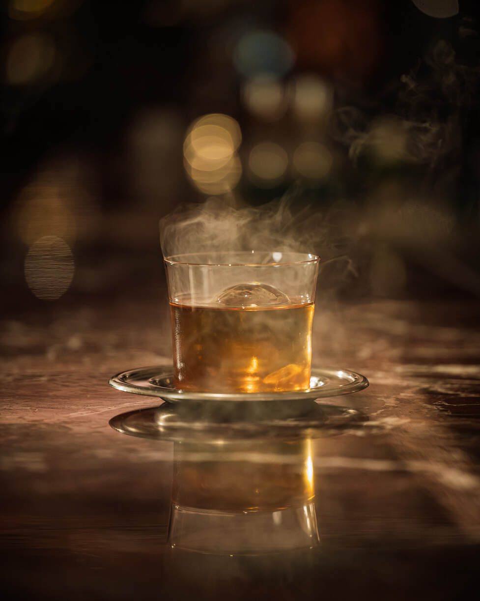 In der renommierten Alcron Bar Prag steht ein dampfendes Glas warmen Whiskys auf einer Untertasse, vor einer reflektierenden Oberfläche mit einem verschwommenen, warm getönten Hintergrund.