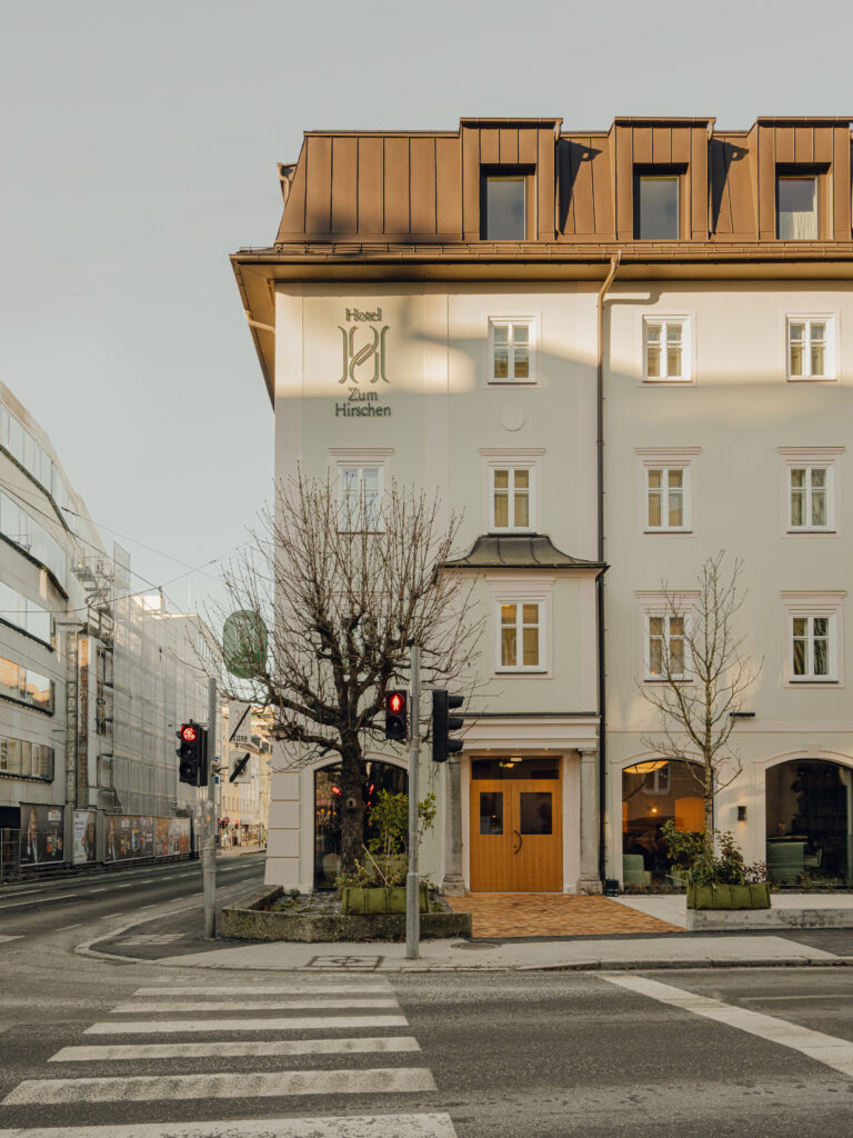 Eckansicht eines beigen Gebäudes mit der Aufschrift „Hotel Zum Hirschen“, gekrönt von einem braunen Dach. Davor stehen zwei Bäume, und ein Zebrastreifen führt Sie zu einer Ampel.