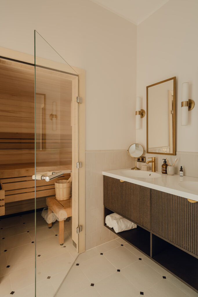 Ein Badezimmer, das an die Eleganz der Alcron Bar Prag erinnert, verfügt über einen Schminktisch mit großem Spiegel, zwei Waschbecken und eine Glastür, die zu einer Sauna mit Holzbänken führt. Die beigefarbenen Wände und der hell geflieste Boden des Zimmers verleihen ihm einen edlen Charme.