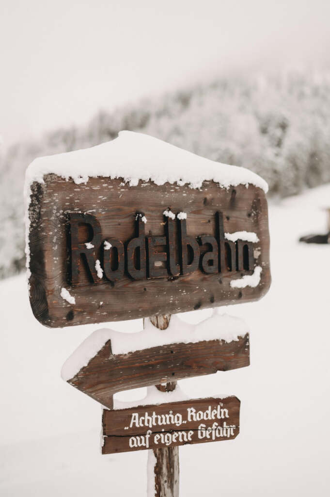 Schneebedecktes Holzschild mit der Aufschrift „Rodelbahn“ und einem Pfeil, begleitet von einer Warnung in deutscher Sprache, dass das Rodeln auf eigene Gefahr erfolgt. Im Hintergrund eine verschneite Landschaft.
