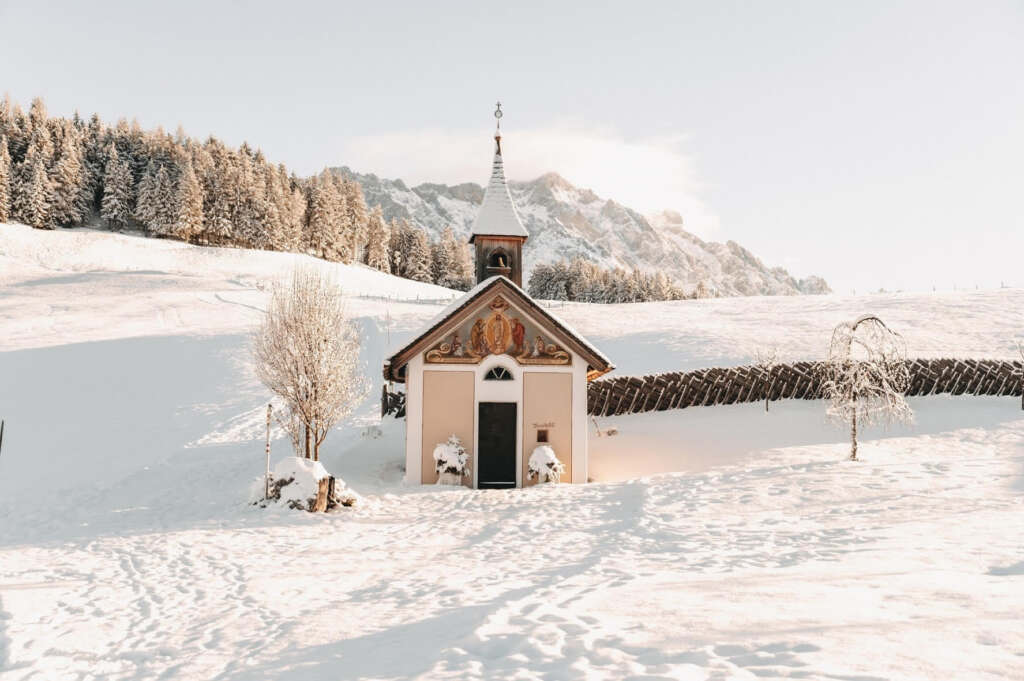 Eine kleine Kapelle steht in einer schneebedeckten Landschaft mit schneebedeckten Bergen und Bäumen im Hintergrund. Die Kapelle hat ein Spitzdach und dekorative Elemente über dem Eingang.