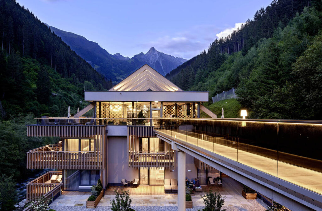 Modernes mehrstöckiges Gebäude mit großen Glasfenstern und Balkonen, gelegen in einem bewaldeten Berggebiet. Eine Brücke verbindet das Gebäude mit einer oberen Etage. Im Hintergrund sind Berge zu sehen.