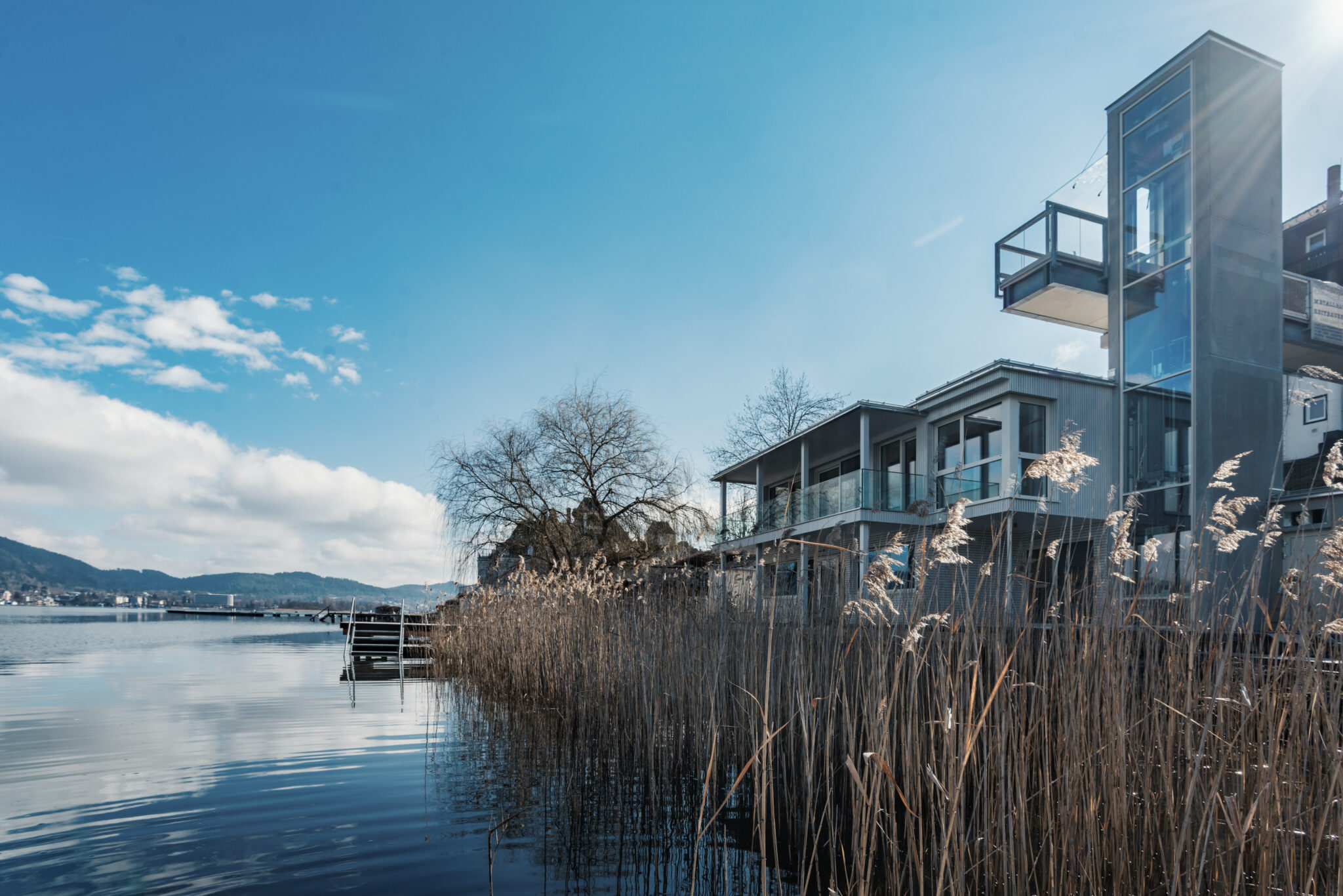 Dieses moderne Haus liegt an einem unberührten See und verfügt über große Fenster und einen Außenaufzug. Hohes Schilf wiegt sich unter einem klaren blauen Himmel, über das Wasser hinweg sind Hügel und eine entfernte Stadt sichtbar – eine idyllische Umgebung, die an die ruhigen Landschaften in der Nähe des Original Mayr Medical Resorts erinnert.