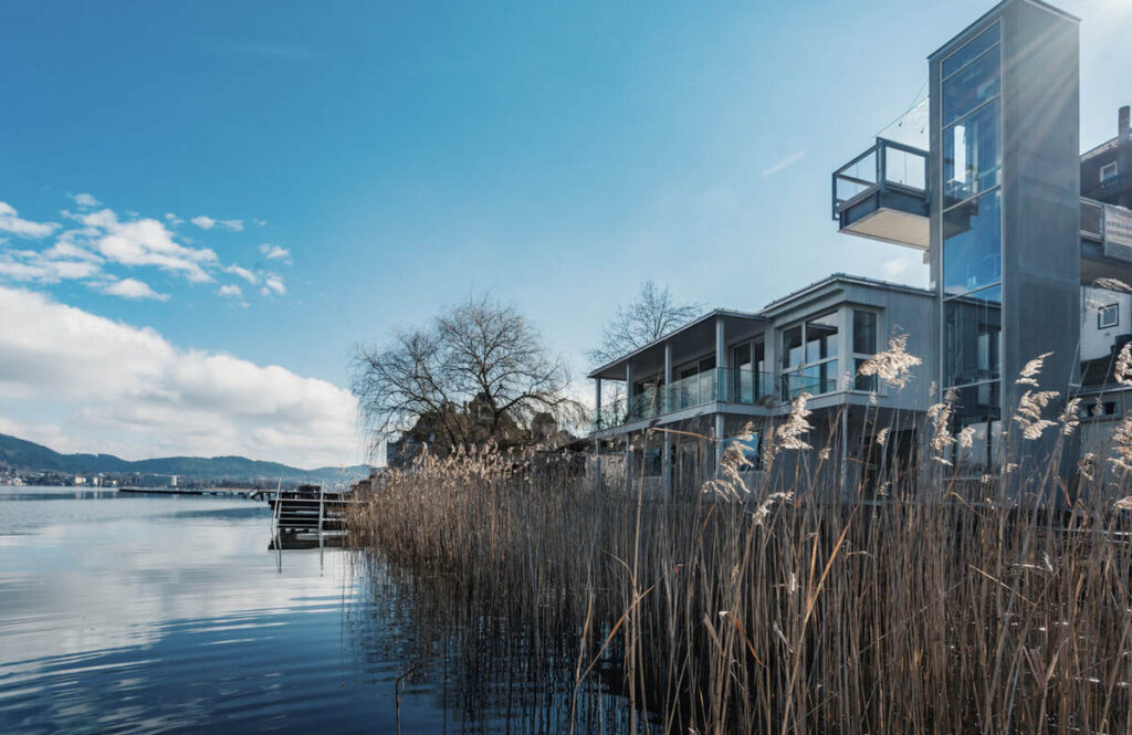Dieses moderne Haus liegt an einem unberührten See und verfügt über große Fenster und einen Außenaufzug. Hohes Schilf wiegt sich unter einem klaren blauen Himmel, über das Wasser hinweg sind Hügel und eine entfernte Stadt sichtbar – eine idyllische Umgebung, die an die ruhigen Landschaften in der Nähe des Original Mayr Medical Resorts erinnert.