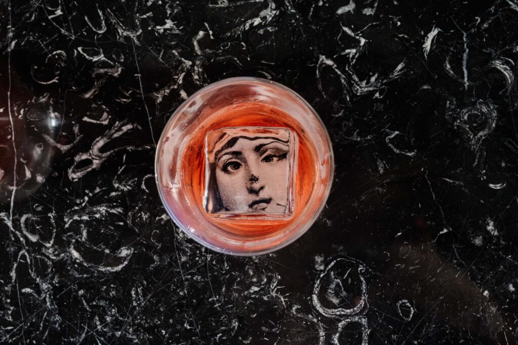 Ein Glas mit einem roten Getränk enthält einen Eiswürfel mit einem schwarz-weißen Porträt eines Gesichts. Das Glas steht auf einer schwarzen Marmoroberfläche.