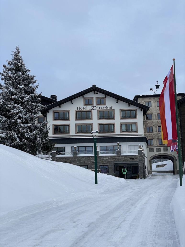 Auffahrt zum Hotel Zürserhof in Österreich. Auf der linken Seite ist eine große, verschneite Tanne und auf der rechten Seite sind verschiedene Fahnen unterschiedlicher Länder zu sehen.