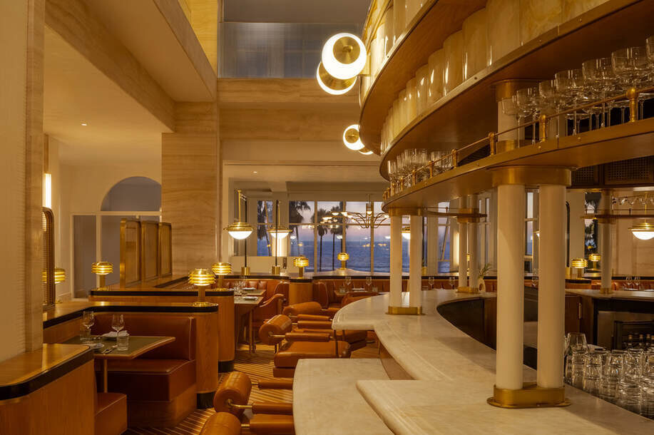 Elegantes Restaurant-Interieur im Art-Deco-Stil, mit warmer Beleuchtung und geschwungenen Sitzgelegenheiten. Große Fenster bieten bei Sonnenuntergang einen Blick auf das Meer.