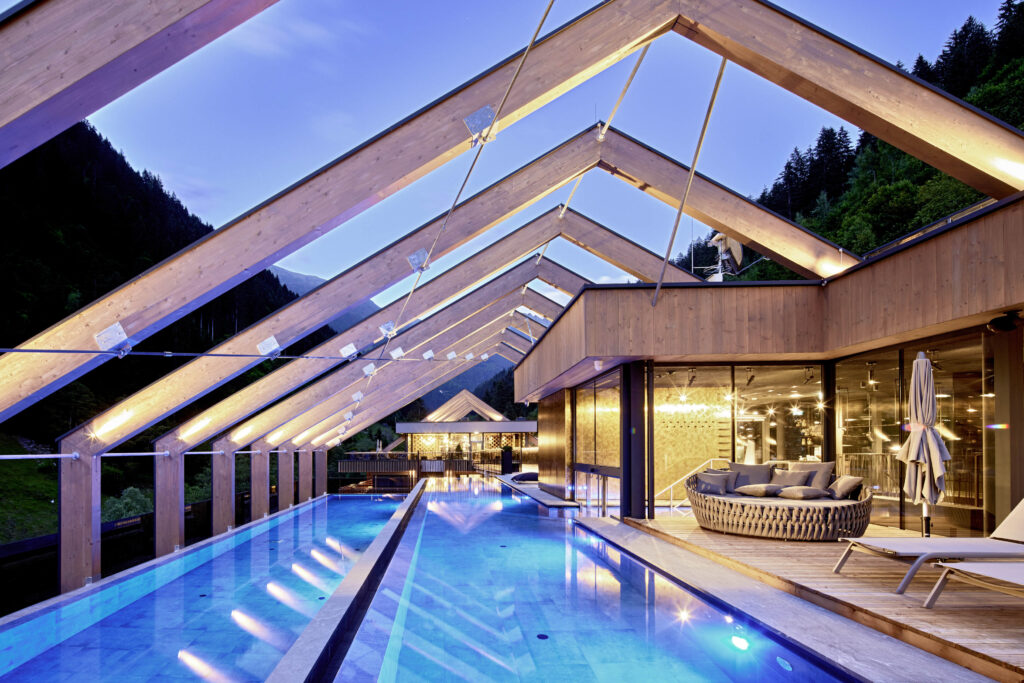 Moderner Innen- und Außenpool mit Dach aus Holzrahmen, der in der Abenddämmerung beleuchtet wird. Die Umgebung umfasst bewaldete Hügel, einen Sitzbereich mit Kissen und große Fenster.