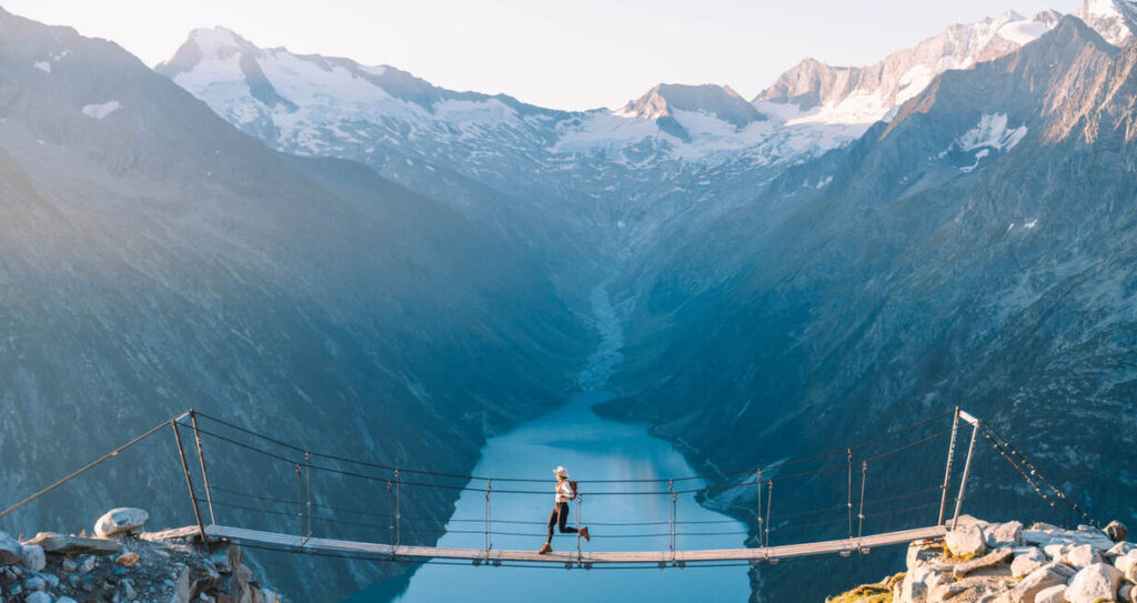 Eine Person läuft über eine Hängebrücke über ein Gebirgstal, darunter fließt ein Fluss, umgeben von schneebedeckten Gipfeln unter einem klaren Himmel.