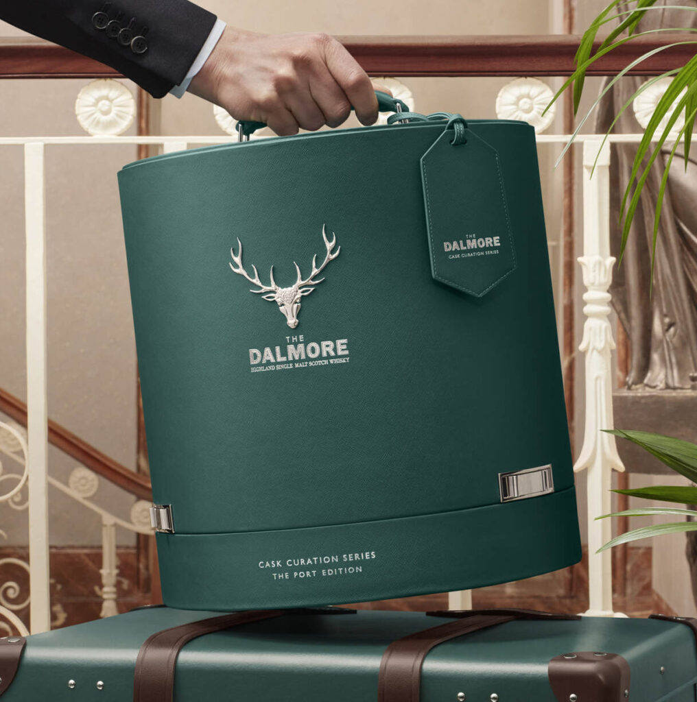 Eine Person hält eine grüne, zylindrische Dalmore-Whisky-Box mit einem silbernen Hirsch-Logo und Text. Die Box ruht auf einem braunen Koffer mit einer Zimmerpflanze und einer Treppe im Hintergrund.