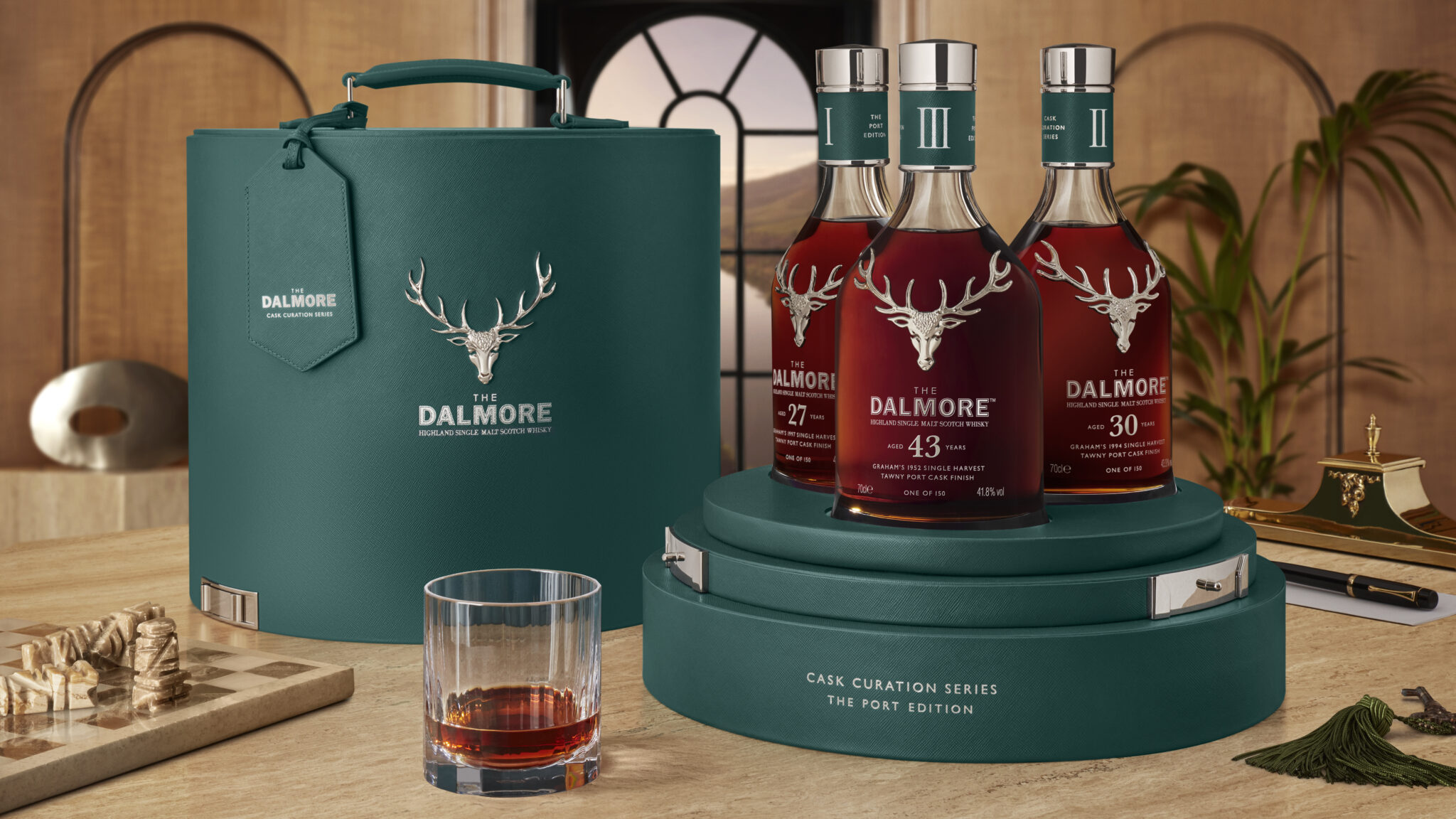 Drei Flaschen The Dalmore Whisky auf einem Tisch mit einem blaugrünen Lederetui, einem Schachspiel und einem gefüllten Glas. Die Etiketten zeigen das Alter von 27, 43 und 30 Jahren an.