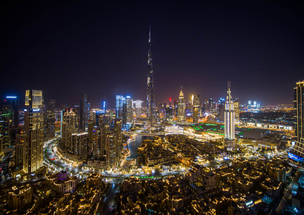 Luftaufnahme einer hell erleuchteten Stadtlandschaft bei Nacht mit einem hohen, beleuchteten Turm in der Mitte. Zahlreiche beleuchtete Gebäude bilden die geschäftige Stadtumgebung.