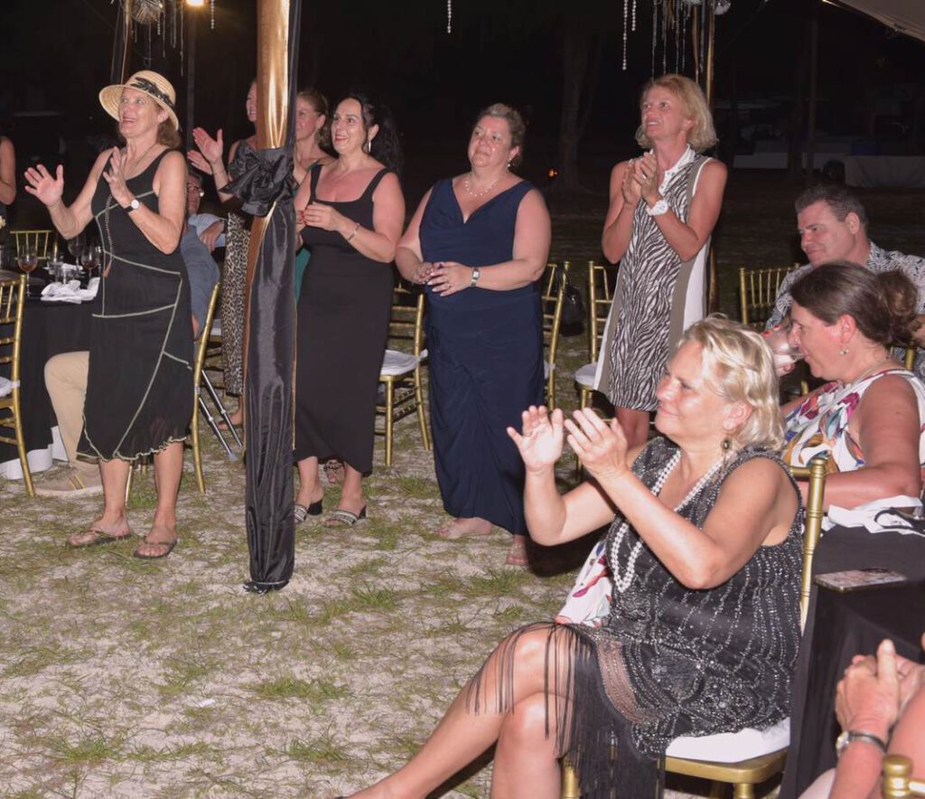 Eine Gruppe von Menschen klatscht und lächelt bei einer Veranstaltung im Freien, im Hintergrund stehen Tische und Stühle.