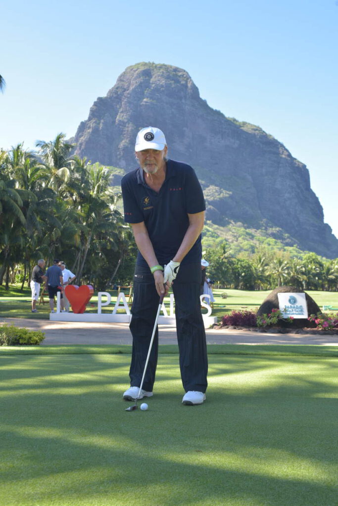 Ein Mann in marineblauem Outfit und Mütze spielt an einem sonnigen Tag Golf mit einem Berg und viel Grün im Hintergrund.