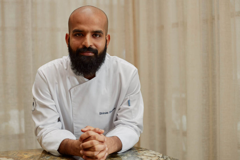 Ein Koch mit Bart und weißer Kochjacke sitzt mit gefalteten Händen an einem Tisch. Im Hintergrund ist ein Vorhang zu sehen.