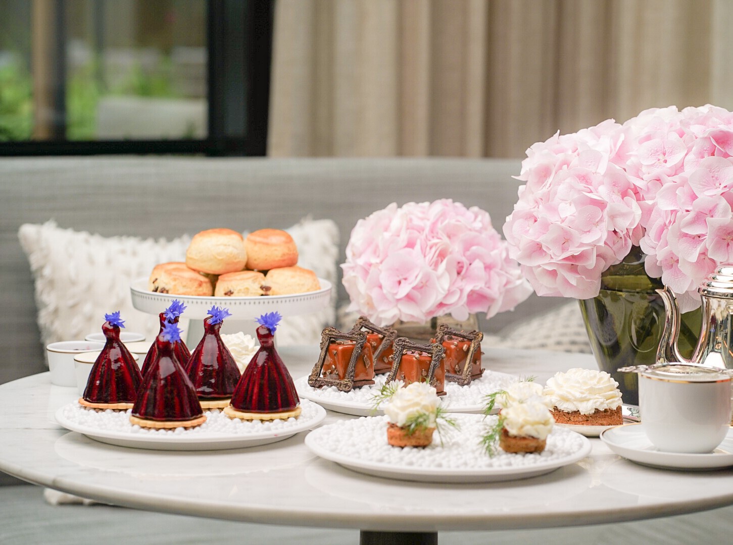 Ein gedeckter Tisch mit verschiedenen Desserts, darunter Gebäck, Torten und Kuchen, neben einer Teekanne und Tassen. Rosa Hortensien schmücken den Tisch.