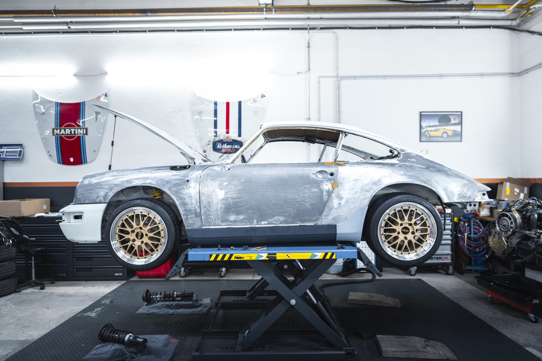 Ein klassischer silberner Sportwagen mit geöffneter Motorhaube steht auf einer Werkstatthebebühne, umgeben von Werkzeugen und Geräten.