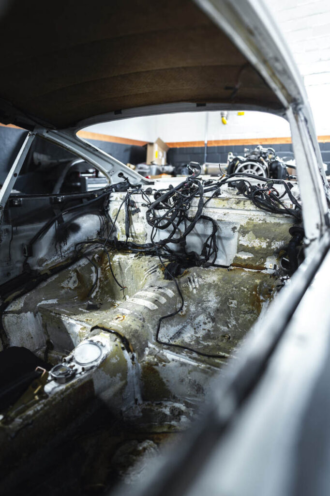 Innenansicht eines zerlegten Autos mit freiliegendem Metall und Kabeln, das in einer Garage geparkt ist, im Hintergrund sind Autoteile zu sehen.
