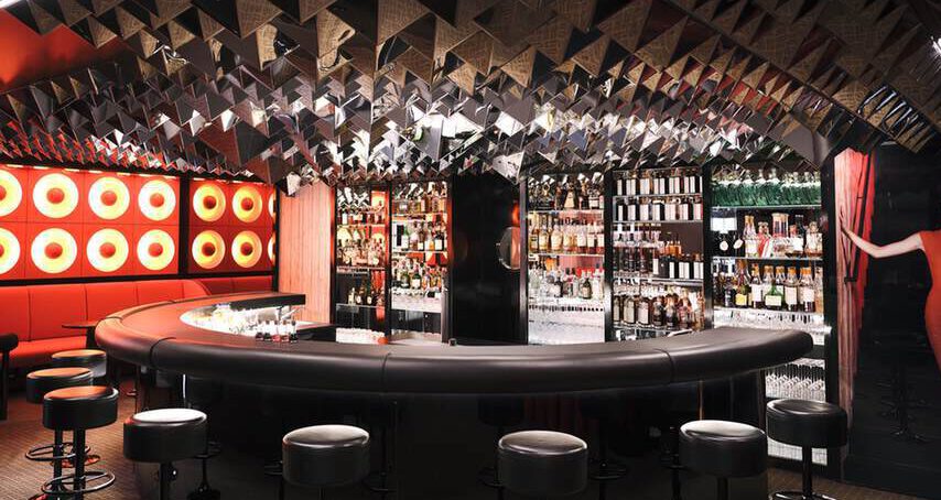 Eine moderne Bar mit geschwungener Theke, umgeben von Hockern. Die Decke ist mit reflektierendem dreieckigem Dekor verziert und an der Wand sind runde rote Lichter zu sehen. An der Seite steht eine Frau in einem roten Kleid.