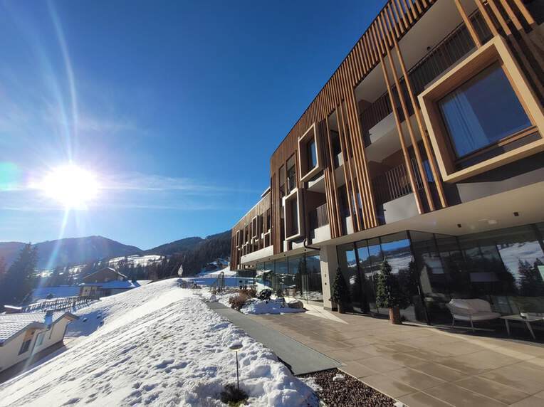 Modernes Gebäude mit Holzakzenten an einem sonnigen Wintertag, umgeben von Schnee und Bergen im Hintergrund.