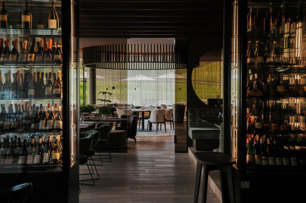 Ein schwach beleuchtetes Restaurant-Interieur mit Weinflaschen auf Glasregalen, dunklen Holzböden, grünen Stühlen und weißen Tischdecken auf runden Tischen in der Nähe großer Fenster.