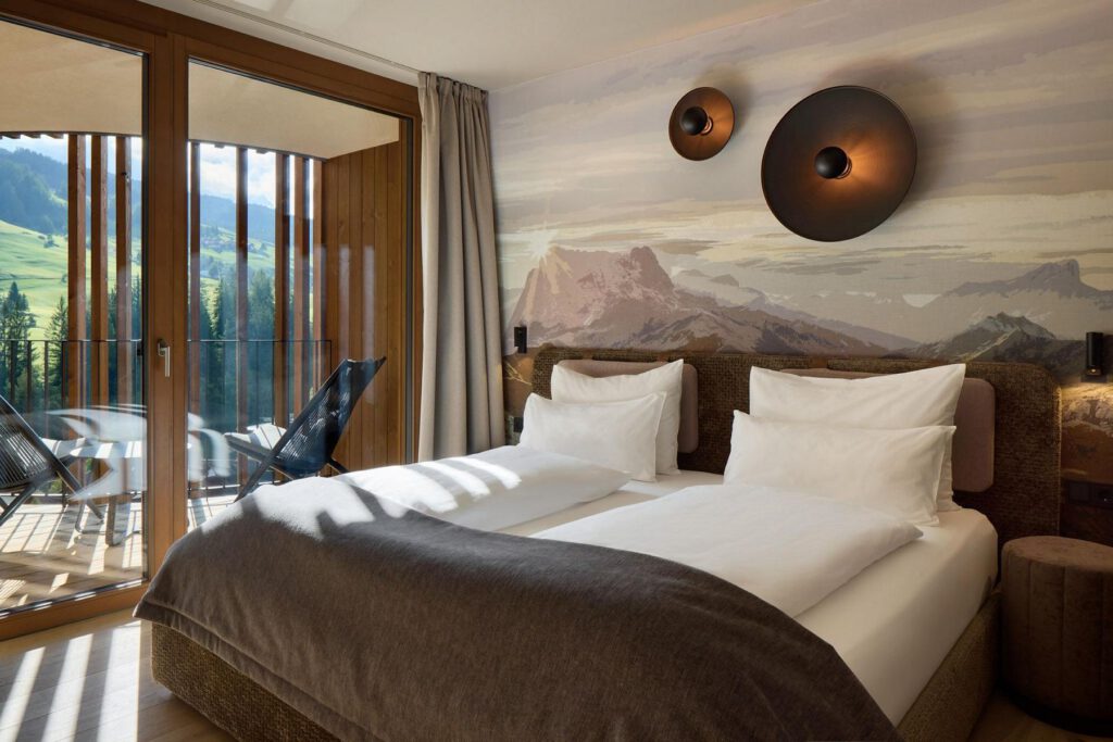 Modernes Hotelzimmer mit zwei Einzelbetten, weißer Bettwäsche und einem Berggemälde an der Wand. Durch die Glastüren ist ein Balkon mit Blick auf Gras und Hügel sichtbar. Über den Betten befinden sich Wandleuchten.