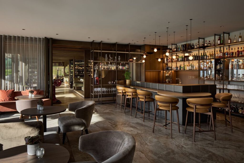 Modernes Bar-Interieur mit eleganter Theke, Barhockern und Regalen voller Flaschen. Runde Tische und gemütliche Stühle füllen den Raum. Warmes Licht und große Fenster unterstreichen das Ambiente.