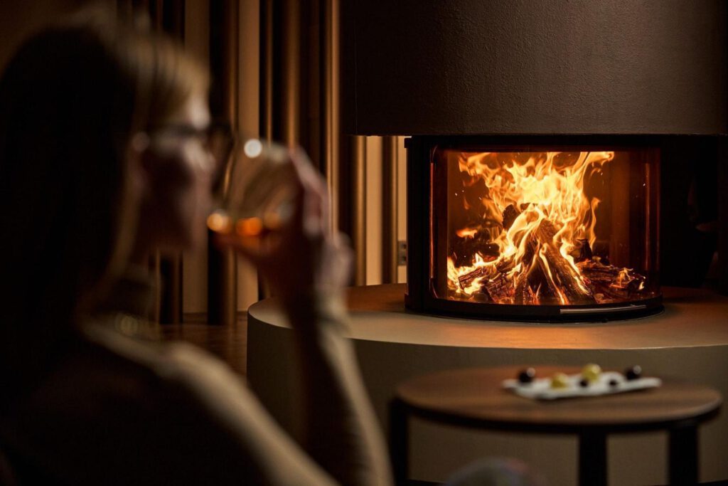 Eine Person sitzt, trinkt aus einem Glas und blickt in einen brennenden Kamin. Im Vordergrund steht ein kleiner Tisch mit einem Teller Essen.