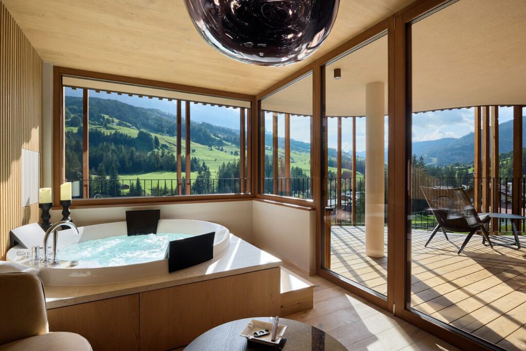 Zimmer mit Whirlpool, großen Fenstern und Blick auf grüne Hügel und blauen Himmel. Draußen gibt es einen Balkon mit Stühlen und eine runde Leuchte hängt von der Holzdecke.
