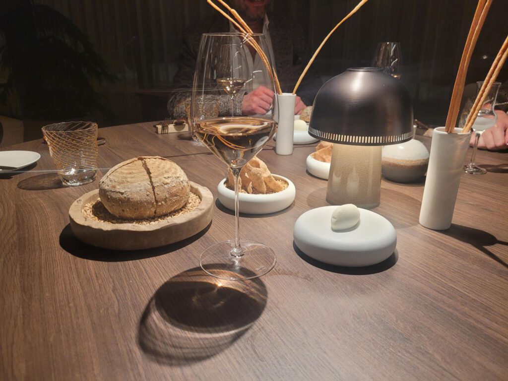 Ein Holztisch mit einem Weinglas, einem Brotlaib, einer kleinen Schüssel Brötchen, einer modernen Lampe und Dekorationsgegenständen. Im Hintergrund sind teilweise Menschen zu sehen.