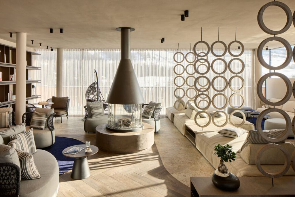 Modernes Wohnzimmer mit rundem Kamin, runden Stühlen und einem ringförmigen Raumteiler. Große Fenster mit transparenten Vorhängen sorgen für Tageslicht. Gepolsterte Sitzgelegenheiten sind rund um den Kamin angeordnet.